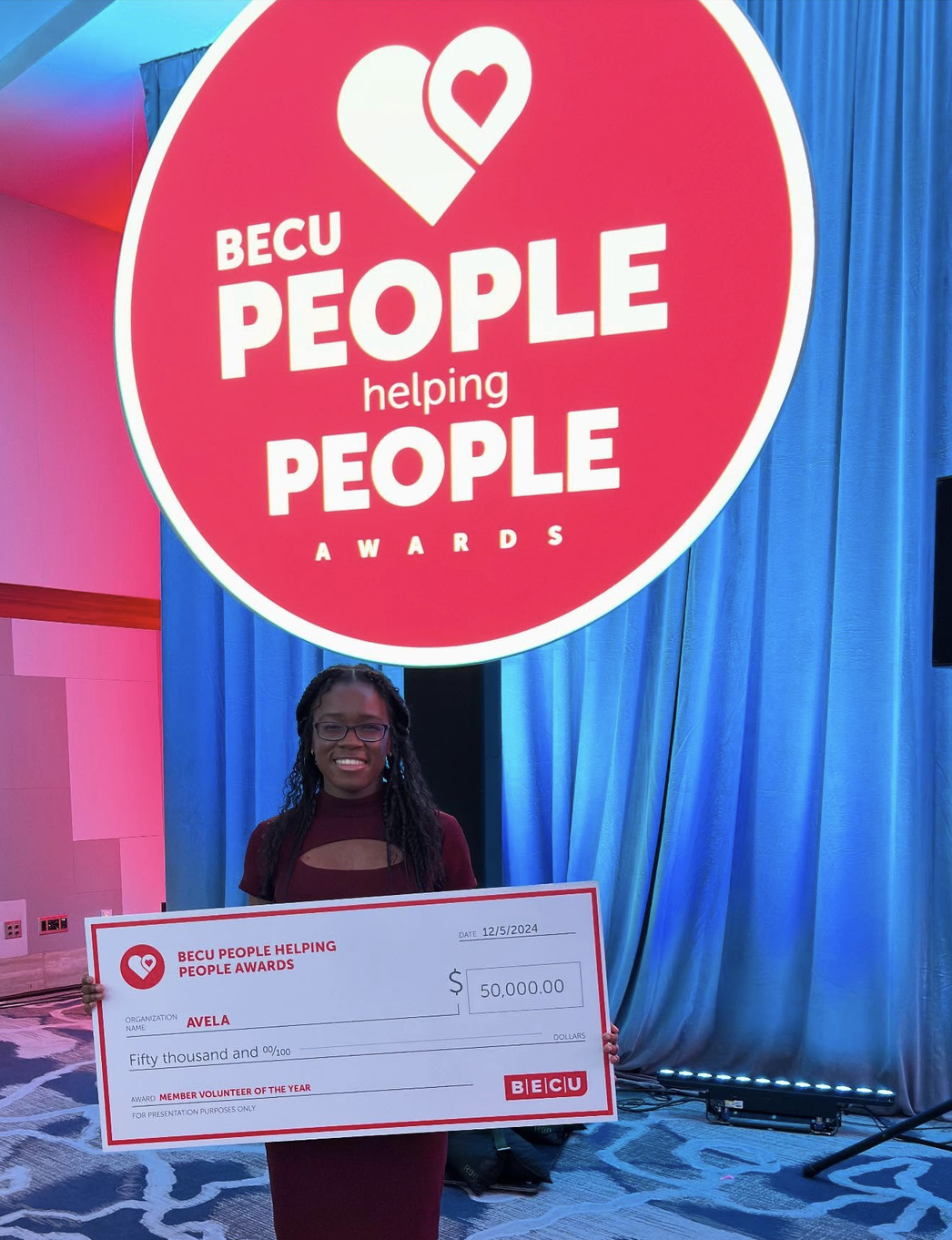 AVELA wins BECU's People Helping People Awards 2024 Ngozi with the 50,000 dollar cheque for AVELA in front of the BECU awards sign.